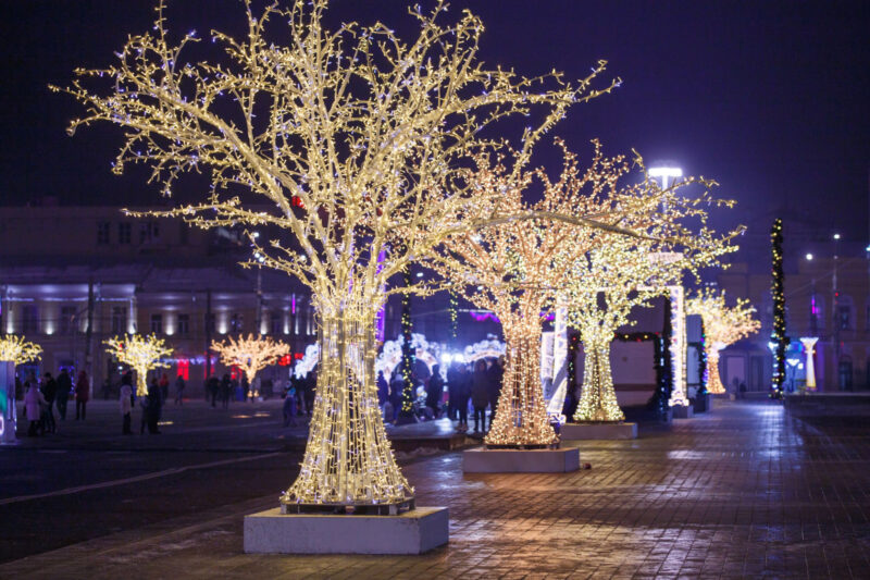 row of luminous decorative trees on night winter street with blurred pedestrians in the background